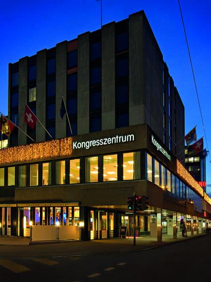 Photo of the Sorell Ador Hotel Bern taken from outside. The sky is dark blue. One can see Swiss and Bern flags as well as the word "Kongresszentrum" on the wall of the building. Copyright Sorell Ador Hotel.