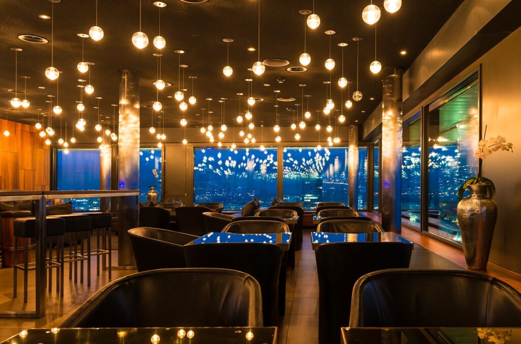 Indoor view of the Skylounge, Zug. One can see a warm room with comfy chairs and many lights. In the background, the windows allow to see buildings.